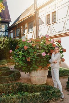 Attractive curly blonde woman walk on the city park street. Girl wear purple hoodie look happy and smiles. Woman make here me gesture standing near pink blooming bush flowers. Happy laughing girl