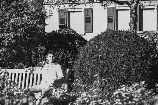 a teenager sits on a bench in the park drinks coffee from a thermo mug and looks into a phone. Portrait of handsome cheerful guy sitting on bench fresh air using device browsing media smm drinking latte urban outside outdoor.