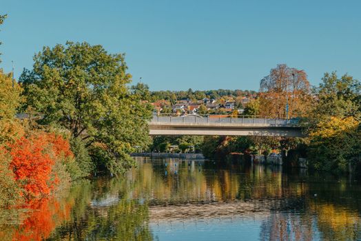 Beautiful small river with clean and clear water front of colorful autumn trees and small old town on the hill agaist nice blue and clouds sky during autumn in Europe.