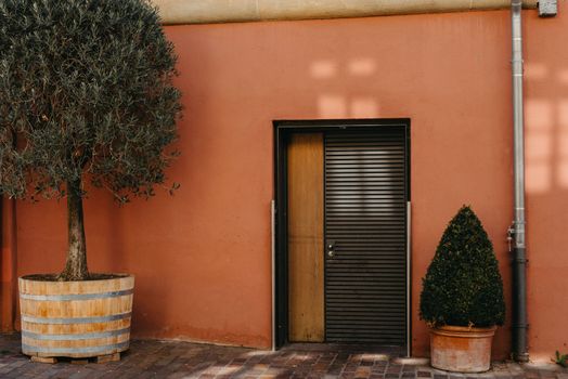 Facade of a modern house with a gray front door and potted flowers. Old historic Italian architecture. Traditional European old town building. Blue wooden door, balcony, blooming bush, pastel walls with sunlight shadows. Aesthetic summer vacation travel background