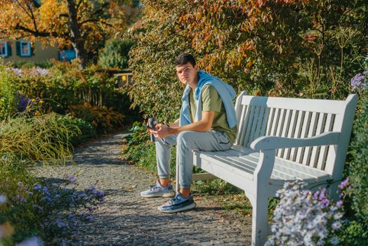 a teenager sits on a bench in the park drinks coffee from a thermo mug and looks into a phone. Portrait of handsome cheerful guy sitting on bench fresh air using device browsing media smm drinking latte urban outside outdoor.