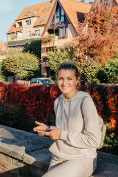 Young fashionable teenage girl with smartphone in park in autumn sitting at smiling. Trendy young woman in fall in park texting. Retouched, vibrant colors. Beautiful blonde teenage girl wearing casual modern autumn outfit sitting in park in autumn. Retouched, vibrant colors, brownish tones.