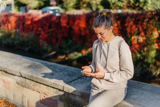 Young fashionable teenage girl with smartphone in park in autumn sitting at smiling. Trendy young woman in fall in park texting. Retouched, vibrant colors. Beautiful blonde teenage girl wearing casual modern autumn outfit sitting in park in autumn. Retouched, vibrant colors, brownish tones.