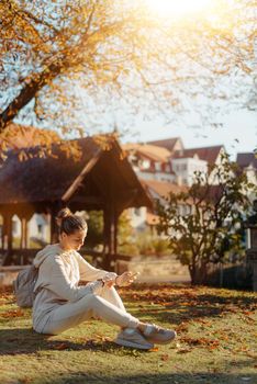 Young fashionable teenage girl with smartphone in park in autumn sitting at smiling. Trendy young woman in fall in park texting. Retouched, vibrant colors. Beautiful blonde teenage girl wearing casual modern autumn outfit sitting in park in autumn. Retouched, vibrant colors, brownish tones.