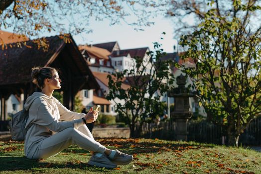 Young fashionable teenage girl with smartphone in park in autumn sitting at smiling. Trendy young woman in fall in park texting. Retouched, vibrant colors. Beautiful blonde teenage girl wearing casual modern autumn outfit sitting in park in autumn. Retouched, vibrant colors, brownish tones.