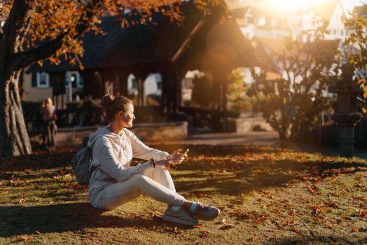 Young fashionable teenage girl with smartphone in park in autumn sitting at smiling. Trendy young woman in fall in park texting. Retouched, vibrant colors. Beautiful blonde teenage girl wearing casual modern autumn outfit sitting in park in autumn. Retouched, vibrant colors, brownish tones.