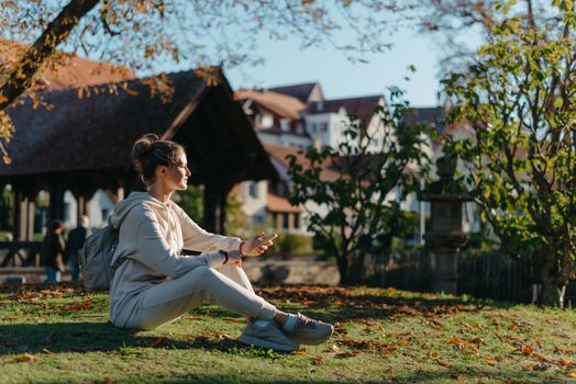 Young fashionable teenage girl with smartphone in park in autumn sitting at smiling. Trendy young woman in fall in park texting. Retouched, vibrant colors. Beautiful blonde teenage girl wearing casual modern autumn outfit sitting in park in autumn. Retouched, vibrant colors, brownish tones.