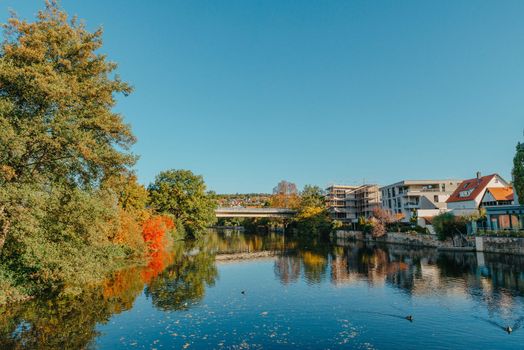 Beautiful small river with clean and clear water front of colorful autumn trees and small old town on the hill agaist nice blue and clouds sky during autumn in Europe.