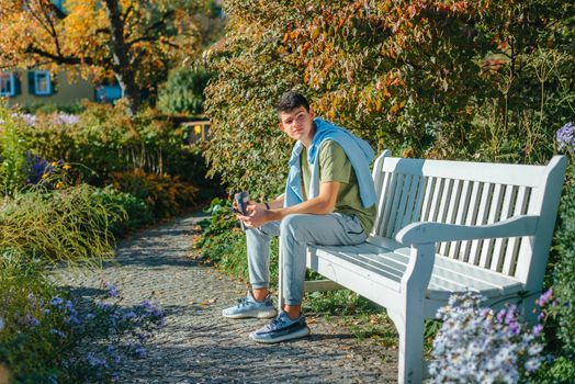 a teenager sits on a bench in the park drinks coffee from a thermo mug and looks into a phone. Portrait of handsome cheerful guy sitting on bench fresh air using device browsing media smm drinking latte urban outside outdoor.