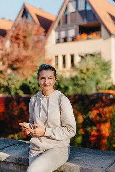 Young fashionable teenage girl with smartphone in park in autumn sitting at smiling. Trendy young woman in fall in park texting. Retouched, vibrant colors. Beautiful blonde teenage girl wearing casual modern autumn outfit sitting in park in autumn. Retouched, vibrant colors, brownish tones.