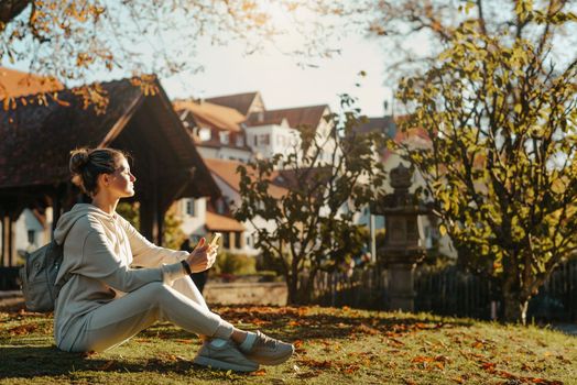 Young fashionable teenage girl with smartphone in park in autumn sitting at smiling. Trendy young woman in fall in park texting. Retouched, vibrant colors. Beautiful blonde teenage girl wearing casual modern autumn outfit sitting in park in autumn. Retouched, vibrant colors, brownish tones.