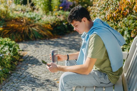 a teenager sits on a bench in the park drinks coffee from a thermo mug and looks into a phone. Portrait of handsome cheerful guy sitting on bench fresh air using device browsing media smm drinking latte urban outside outdoor.