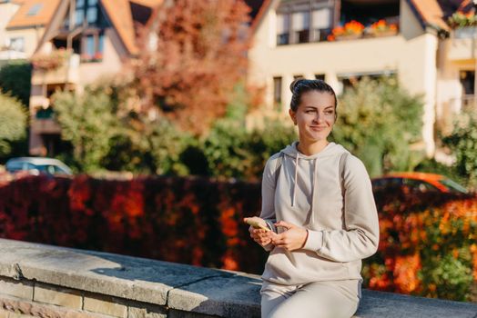 Young fashionable teenage girl with smartphone in park in autumn sitting at smiling. Trendy young woman in fall in park texting. Retouched, vibrant colors. Beautiful blonde teenage girl wearing casual modern autumn outfit sitting in park in autumn. Retouched, vibrant colors, brownish tones.