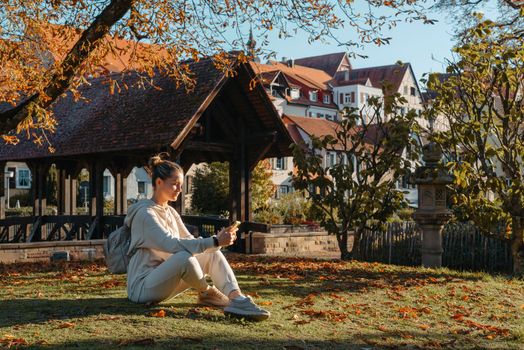 Young fashionable teenage girl with smartphone in park in autumn sitting at smiling. Trendy young woman in fall in park texting. Retouched, vibrant colors. Beautiful blonde teenage girl wearing casual modern autumn outfit sitting in park in autumn. Retouched, vibrant colors, brownish tones.