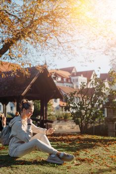 Young fashionable teenage girl with smartphone in park in autumn sitting at smiling. Trendy young woman in fall in park texting. Retouched, vibrant colors. Beautiful blonde teenage girl wearing casual modern autumn outfit sitting in park in autumn. Retouched, vibrant colors, brownish tones.