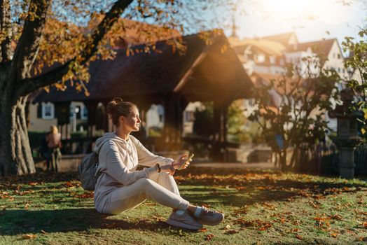 Young fashionable teenage girl with smartphone in park in autumn sitting at smiling. Trendy young woman in fall in park texting. Retouched, vibrant colors. Beautiful blonde teenage girl wearing casual modern autumn outfit sitting in park in autumn. Retouched, vibrant colors, brownish tones.