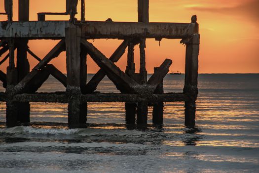 Looking out at Broken old structure remains of pier in the sea. Small wave crashing into the textured rusty pier posts in the during beautiful twilight in the sea. No focus, specifically.