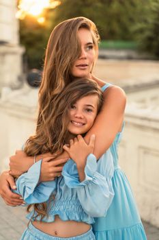 Mother aughter sunset. in blue dresses with flowing long hair against the backdrop of sunset. The woman hugs and presses the girl to her