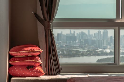 Three red Pillows in the corner on Sofa bed with Curtain near Glass window with city view. Landscaped Resting Area for people who living in lifestyle, Copy space, Selective focus.