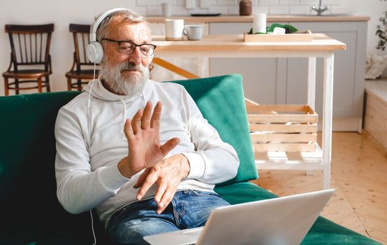 Elderly man making video call on laptop in room waving to screen and chatting with children - modern technologies communication internet