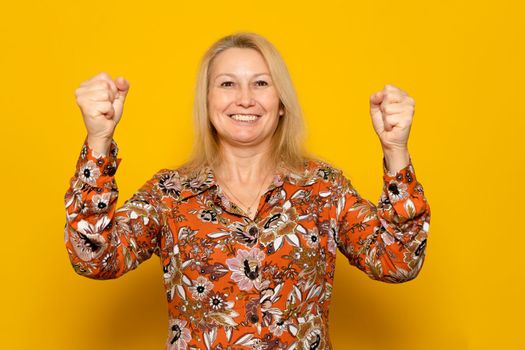 Happy caucasian successful woman in a patterned dress with raised hands smiling and celebrating success over yellow background
