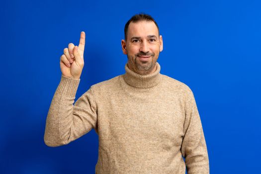 Insightful intelligent proactive man hispanic man 40s wearing turtleneck holding index finger up with great new idea isolated on blue background studio portrait. People lifestyle concept