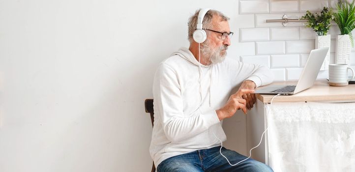 Handsome old man dressed in hoodie and eyeglasses is using laptop and smiling while listen music sitting on couch home