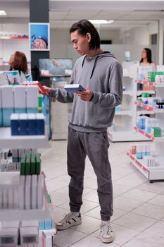 Young asian client examining supplements on shelves, man looking at pills boxes in drugstore filled with health care products like vitamins and drugs. Searching for medical supplies.