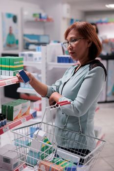 Asian customer looking at prescription drugs box to buy disease treatment and medicinal supplements. Woman reading leaflet of medicaments package, drugstore shelves with medical supplies.