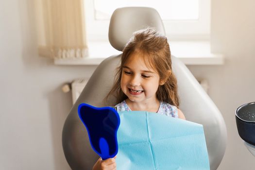 Child looking in the mirror at the dentist. Happy child patient of dentistry. Teeth treatment. Attractive kid girl sitting in dental office and smiling