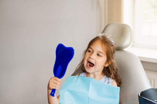 Child looking in the mirror at the dentist. Happy child patient of dentistry. Teeth treatment. Attractive kid girl sitting in dental office and smiling