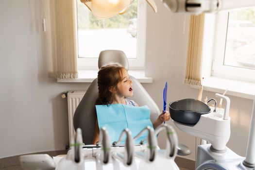 Teeth treatment. Child looking in the mirror at the dentist. Happy child patient of dentistry. Attractive kid girl sitting in dental office and smiling
