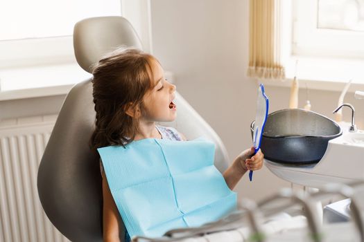 Child looking in the mirror at the dentist. Happy child patient of dentistry. Teeth treatment. Attractive kid girl sitting in dental office and smiling