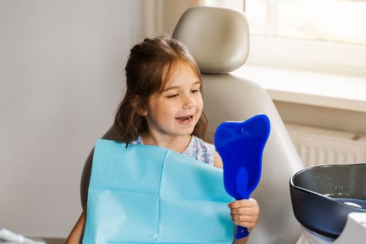 Child looking in the mirror at the dentist. Happy child patient of dentistry. Teeth treatment. Attractive kid girl sitting in dental office and smiling