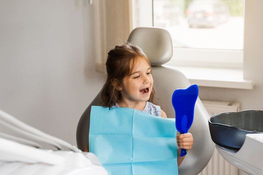 Teeth treatment. Child looking in the mirror at the dentist. Happy child patient of dentistry. Attractive kid girl sitting in dental office and smiling