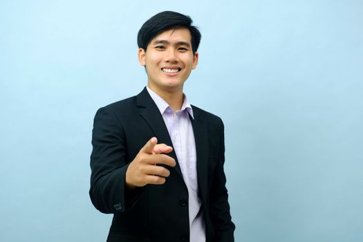 Portriat of asian young smart happy businessman dressed in suit standing straight, smiling, and pointing finger at camera with isolated light blue background. Business concept.