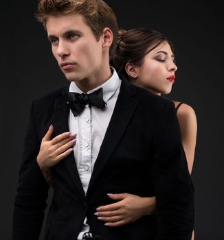 Sexy gentleman in a siut and bow-tie with his girl-friend embracing him cropped isolated shot on black background