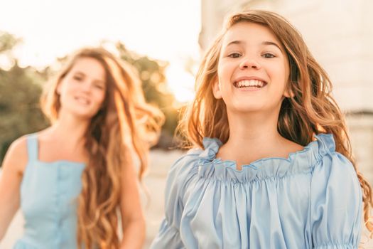 Mother daughter sunset. in blue dresses with flowing long hair against the backdrop of a sunset and a white building. They run and smile at the camera. Family stories on the weekend.