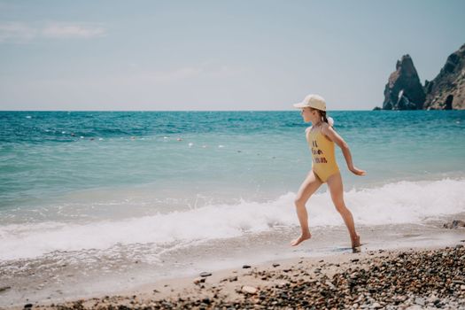 Cute little girl running along the seashore against a clear blue sea and rejoices in the rays of the summer sun. Beautiful girl in yellow swimsuit running and having fun on tropical beach