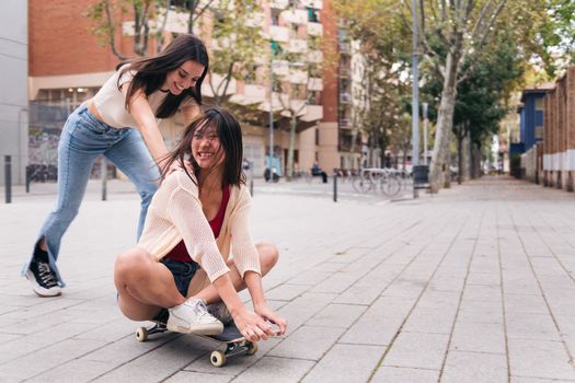 two young women laughing and having a good time playing with a skateboard down a city street, concept of friendship and teenager lifestyle, copy space for text