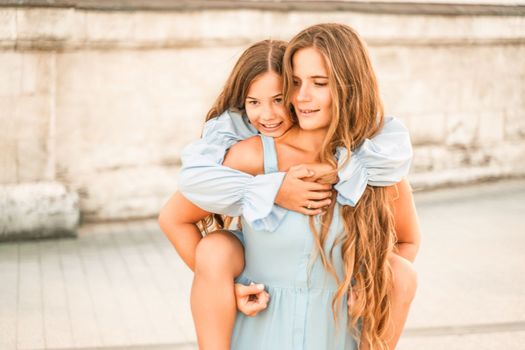 Mother of the daughter walks playing. Mother holds the girl on her back, holding her legs, and her daughter hugs her by the shoulders. Dressed in blue dresses