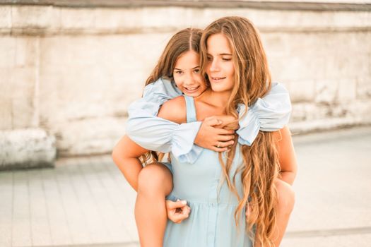 Mother of the daughter walks playing. Mother holds the girl on her back, holding her legs, and her daughter hugs her by the shoulders. Dressed in blue dresses