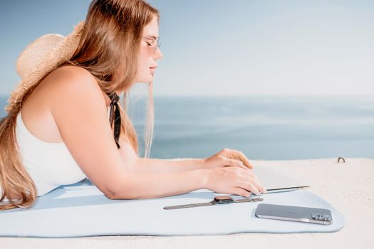 Successful business woman in yellow hat working on laptop by the sea. Pretty lady typing on computer at summer day outdoors. Freelance, travel and holidays concept.
