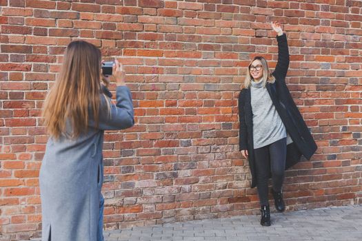 Girl takes picture of her female friend in front of brick wall in urban street - photographer and youth urban lifestyle