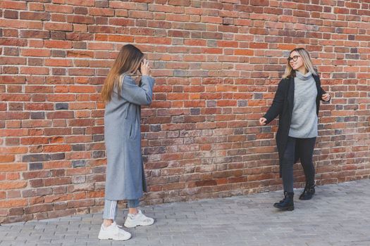 Girl takes picture of her female friend in front of brick wall