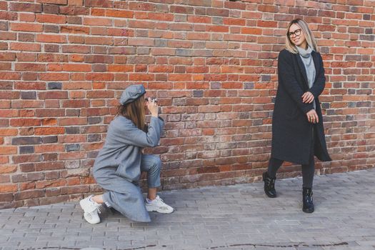 Girl takes picture of her female friend in front of brick wall