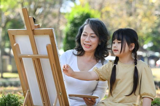 Happy middle aged grandma painting picture with lovely little grandchild, enjoying leisure weekend activity together outdoor.