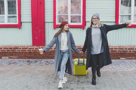 Two cheerful tourist women smiling and walking with suitcases on city street in autumn or spring time - travel and vacation