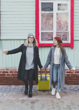 Two cheerful tourist women smiling and walking with suitcases on city street in autumn or spring time - travel and vacation