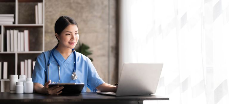 Asian doctor young beautiful woman smiling using working with a laptop computer and her writing something on paperwork or clipboard white paper at hospital desk office, Healthcare medical concept..